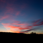 Sonnenuntergang – Unten im Bild die im dunkeln liegende Landschaft, darüber der blauschwarze Himmel mit rotorangen Wolken.