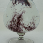 Ein bauchiges Glas mit kurzem Stiel, dass fast bis oben mit Wasser gefüllt ist. Im Wasser hat es Spuren von schwarzer Tinte, die sich durch das klare Wasser ziehen.